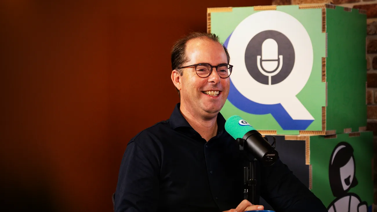 Gast lacht achter de microfoon, met het logo van de podcast achter zich